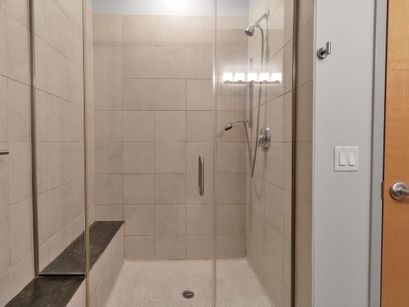A white tiled shower with a glass door and a bench. at The Granite Works Apartments , Buffalo, NY