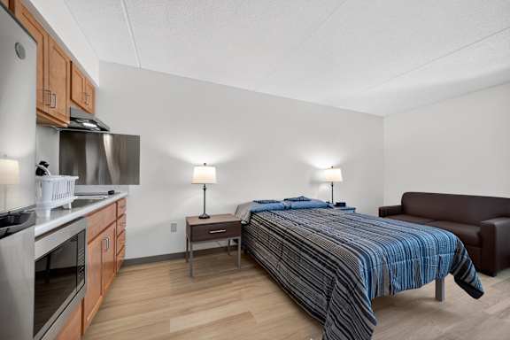 A modern bedroom with a large bed and a kitchen area in the background.