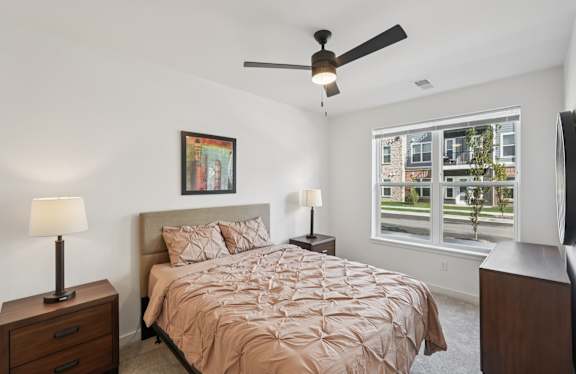 A bedroom with a bed, two lamps, a ceiling fan, and a window overlooking a street.