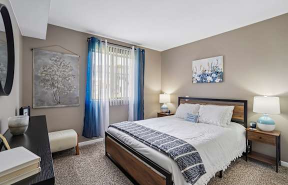 bedroom with tan walls, tan carpeting, and tan headboard. The bed is made in a blue patterned bedding with throw pillows. There are white closet doors to the right of the bed.  