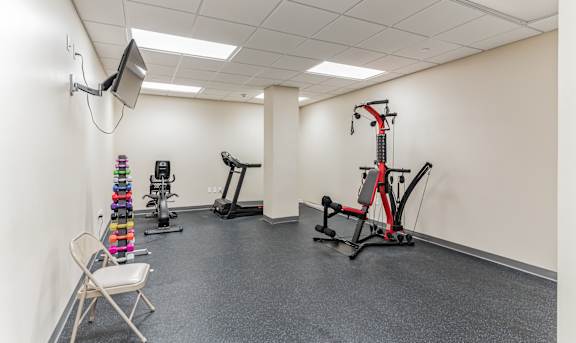 A gym room with a variety of equipment including a treadmill, a stationary bike, a rowing machine, and a chair at 13 State Street Apartments in Schenectady, NY