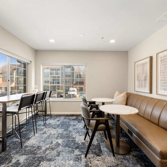 A room with a brown leather couch, a table with chairs, and two framed pictures on the wall.