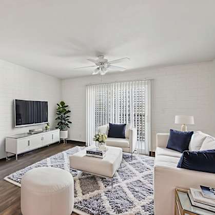 A living room with a white couch, a white chair, a white ottoman, a white rug, a white ceiling fan, a white door, a white cabinet, a white television, and a white wall.