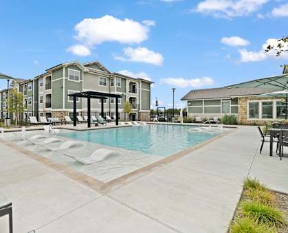 A large swimming pool surrounded by lounge chairs and umbrellas in front of a multi-story apartment building.