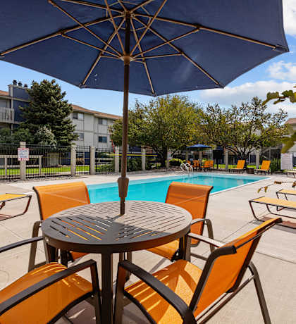 A table with chairs and an umbrella is in the foreground of a pool area.