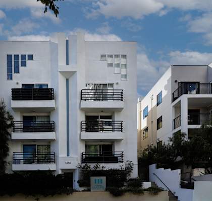 A white building with balconies and windows is surrounded by other buildings and trees.