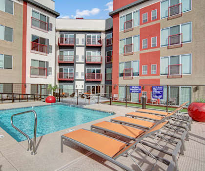 The turquoise outdoor pool with pool chairs, tables, and chairs with the tan and orange apartments in the background at Proxy 333 Apartment Homes. 