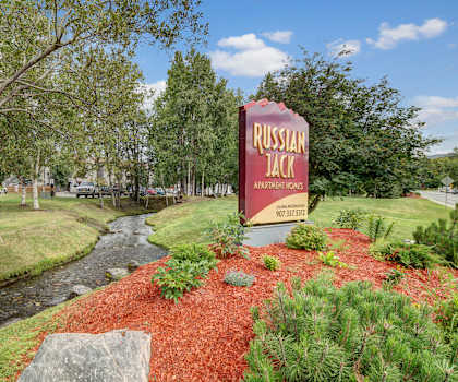 The exterior sign with trees and surrounded by grass and greenery with a creek, the parking lot, and the buildings in the background of Russian Jack Apartment Homes.