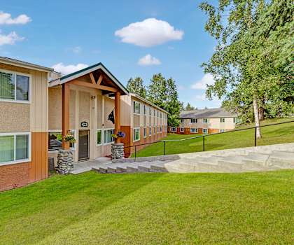 The front entrance to the tan and orange leasing office with white descending marble stairs surrounded by grass and trees at Taiga Apartment Homes. 