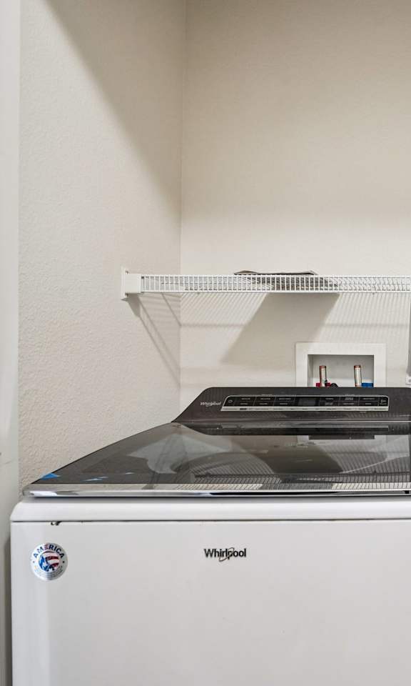 A white Washgood washing machine sits next to a white shelf in a laundry room.