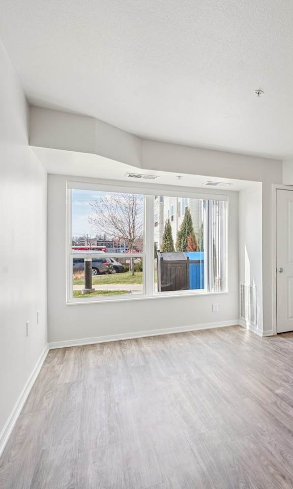 A room with white walls and wooden flooring, with a window showing a view of trees and a blue dumpster.