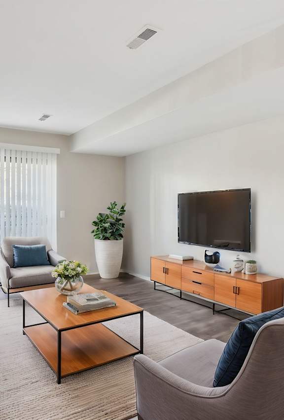 A living room with a brown sofa, a grey armchair, a glass coffee table, a television, and a potted plant at Millennium at Creve Coeur, Creve Coeur, 63141
