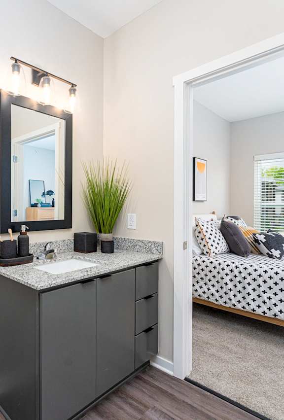 A bathroom with a toilet, sink, mirror, and a bed in the background.