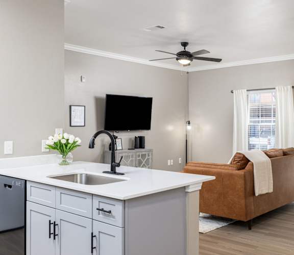 A kitchen with a sink, a fridge, a microwave, a counter, a fan, a sofa, a window, and a vase of flowers. at Clear Creek Meadows Apartments, Texas