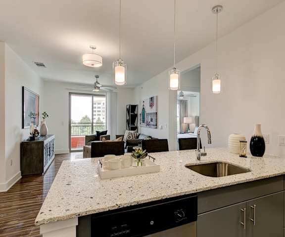 A kitchen with white marble countertops and gray cabinets with a view of the living room with a couch, chairs, coffee table, and doors to the balcony at Florence at the Harbor. 