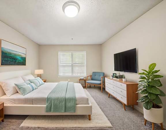 A bedroom with a bed, a chair, a dresser, a lamp, a plant, and a painting on the wall.
