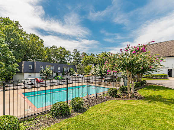 A pool surrounded by a black fence with a tree with pink flowers in the foreground at Pines at Lawrenceville Apartments in Georgia, 30033