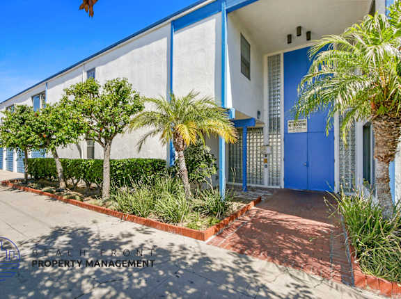 A blue building with a white door and windows, surrounded by greenery and a red brick pathway.