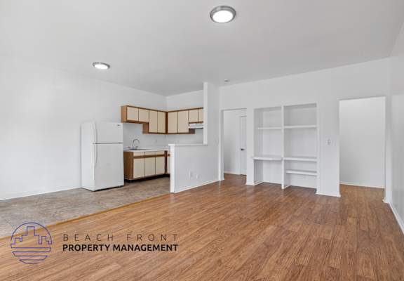 A spacious room with wooden floors and white walls, featuring a kitchenette with a refrigerator, sink, and cabinets.