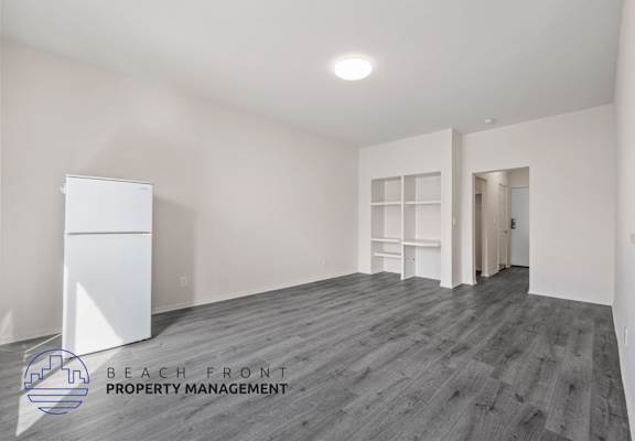 A spacious room with wooden flooring and white walls, with a white refrigerator on the left side.