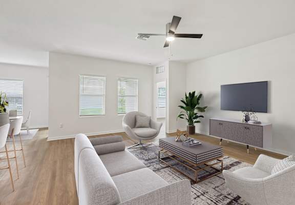 A living room with a grey sofa, a white armchair, a wooden floor, a ceiling fan, and a television on a cabinet.