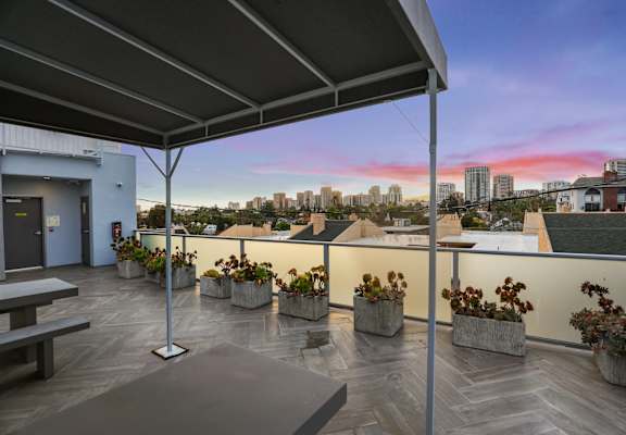 A rooftop patio with a bench, planters, and a view of the city skyline at dusk.