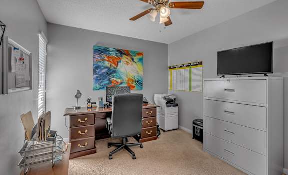 A room with a ceiling fan, a desk with a chair, a filing cabinet, and a television.