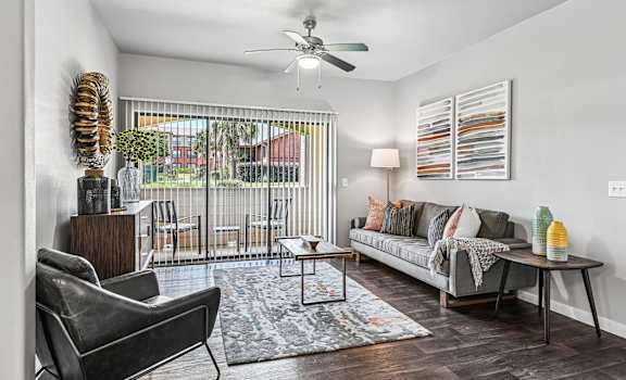 A living room with a grey couch, a black chair, a wooden side table, a floor lamp, a ceiling fan, and a large window with a view of the outside.