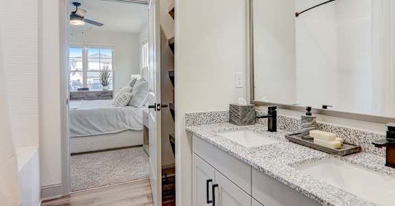 A bathroom with a mirror, sink, and a bedroom with a bed and a window.