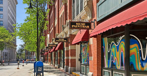 A street with a blue mailbox and a building with a red awning that says "MUSE PAINTRA".