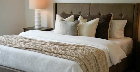 A bed with a white comforter and pillows with a lamp on the side table.