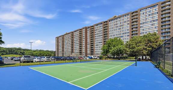 A tennis court with a blue surface and white lines is surrounded by apartment buildings.