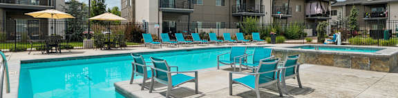 A swimming pool surrounded by lounge chairs and umbrellas in front of apartment buildings.