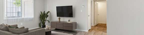 A living room with a grey couch, a brown armchair, a wooden floor, and a flat-screen TV mounted on the wall.