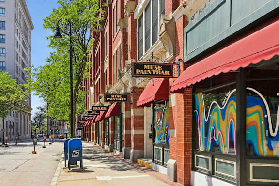 A street with a blue mailbox and a building with a red awning that says "MUSE PAINTRA".