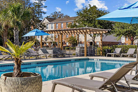 A pool area with a tree in a pot, lounge chairs and umbrellas.