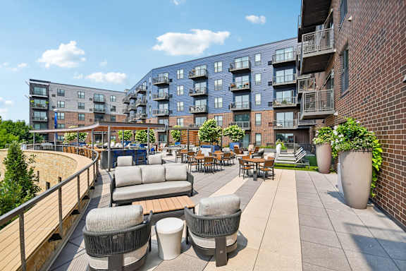 A patio area with couches and tables is surrounded by a brick wall and buildings.
