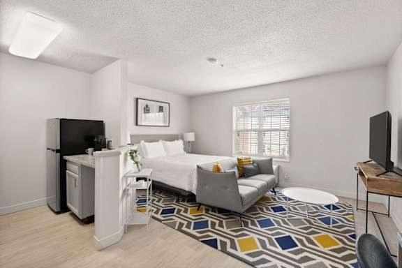 A bedroom with a bed, a chair, a desk, and a fridge.