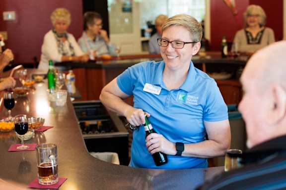 A woman in a blue shirt is pouring a drink for a man at a bar.