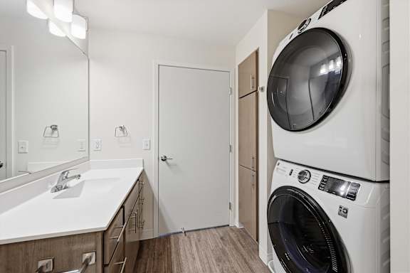 A white washing machine is in a laundry room with a sink and mirror.