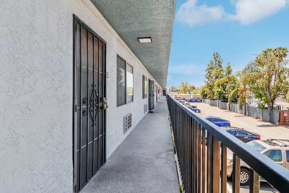 A long, narrow balcony with a metal railing and a white wall with a black gate.