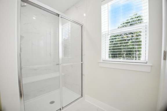 A white bathroom with a glass shower door and a window with blinds.
