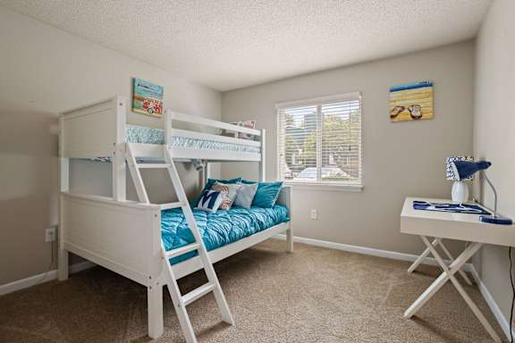 A white bunk bed with a blue mattress and pillows is in a room with a desk and a window.