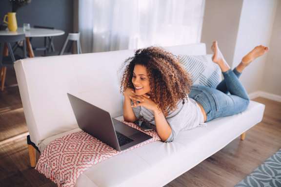 A woman is lying on a couch with her laptop at Heights at Black Bear Apartments, West Virginia, 26501