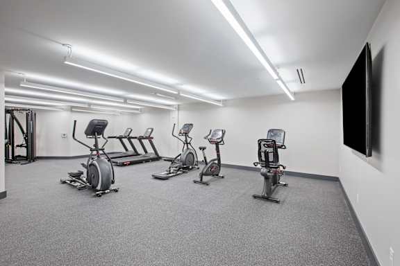 A gym with a grey carpet and a white wall with a television.