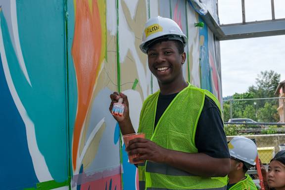 A man in a white helmet and green vest is painting a mural.