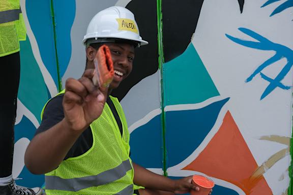 A man in a white helmet and yellow vest is painting a mural.