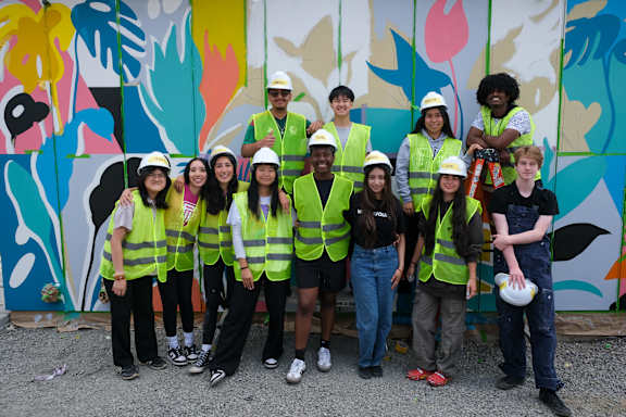 A group of people wearing safety vests and hard hats stand in front of a colorful mural.