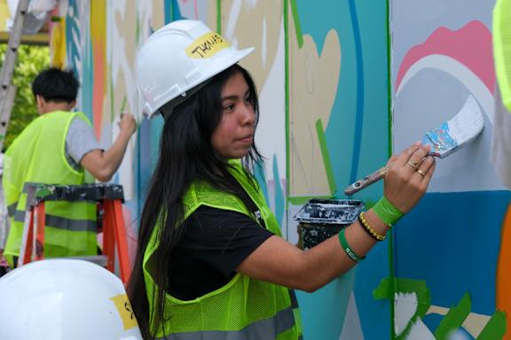 A woman in a white helmet and green vest is painting a mural.