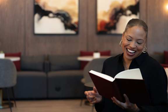 A woman is reading a book in a room with a grey couch and a painting on the wall. at SMB Hollywood Apartments, Los Angeles, CA, 90038